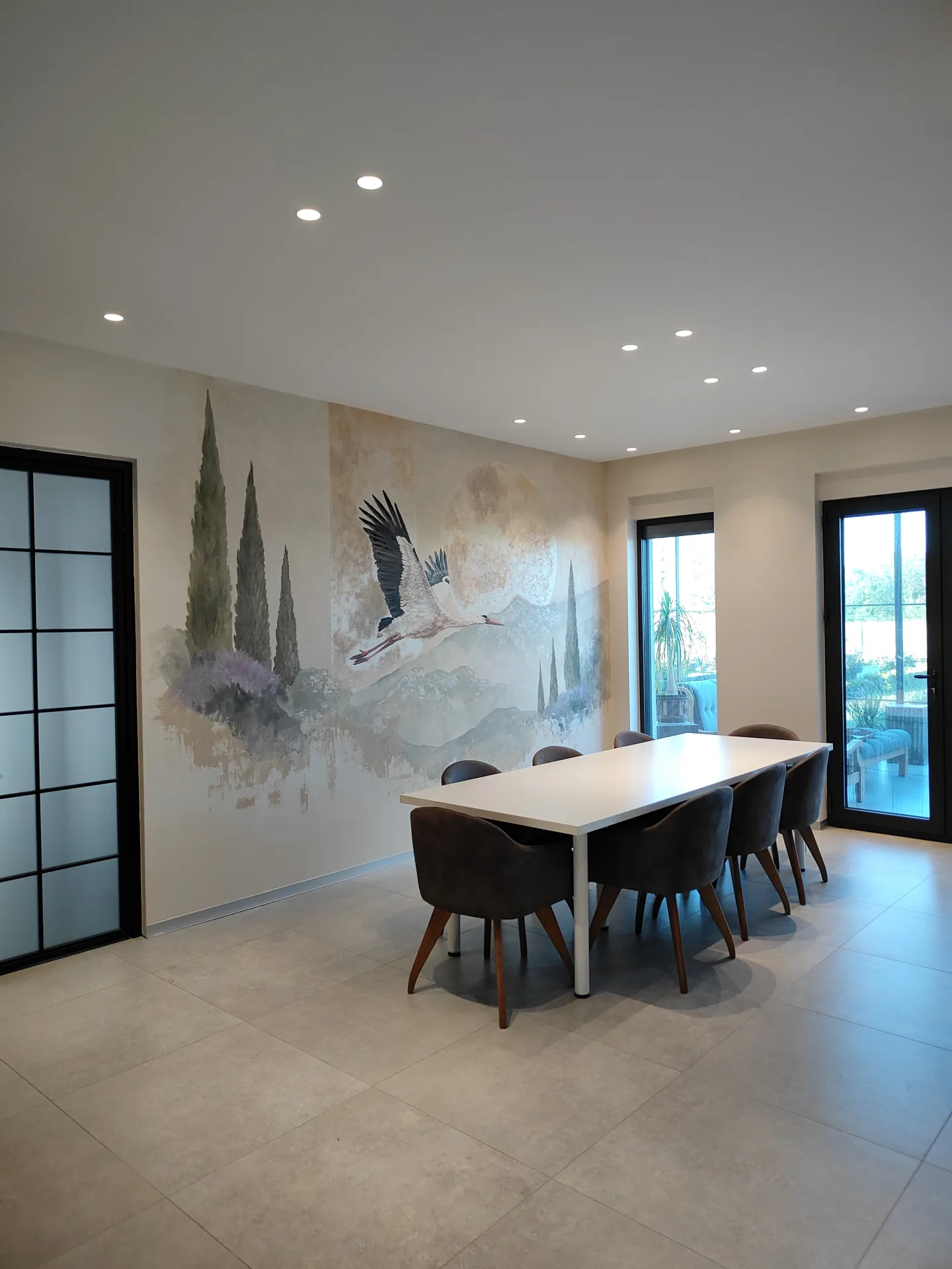Stork in flight above a dining table, with painted trees and hills.